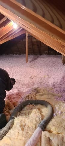 Attic with insulation. A person is working with a hose amid pink and yellow insulation. Wooden beams are visible.