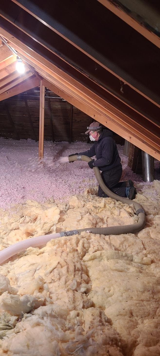 Person installing insulation in an attic. Light-colored insulation is visible, with a hose.