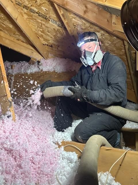 Person in attic with respirator and hose blowing insulation. Pink and white insulation in a wooden attic.