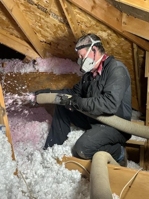 Man in a mask installing pink insulation in an attic, kneeling and holding a hose.