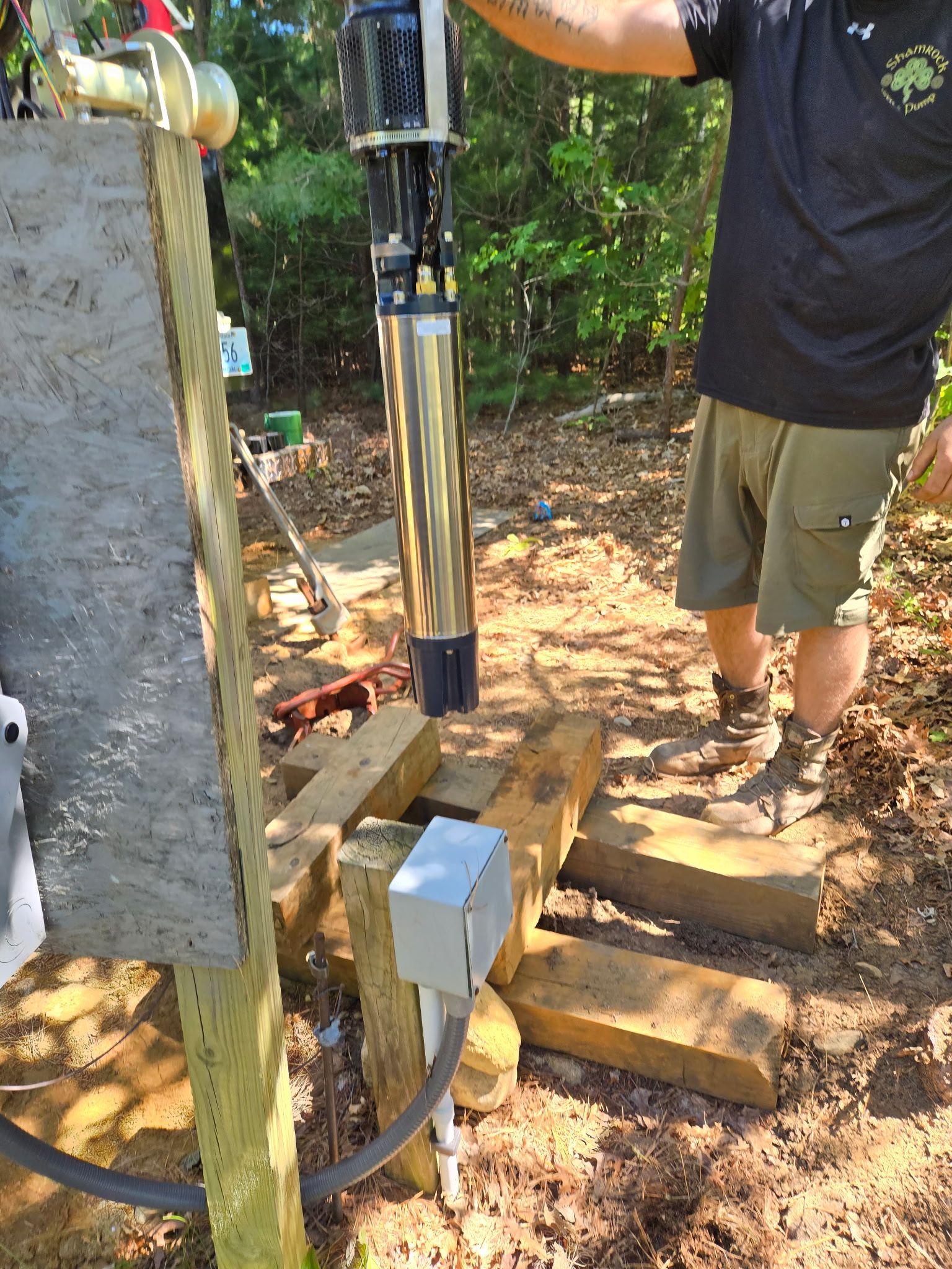 Person installing a submersible water pump next to a wooden electrical box in an outdoor setting.