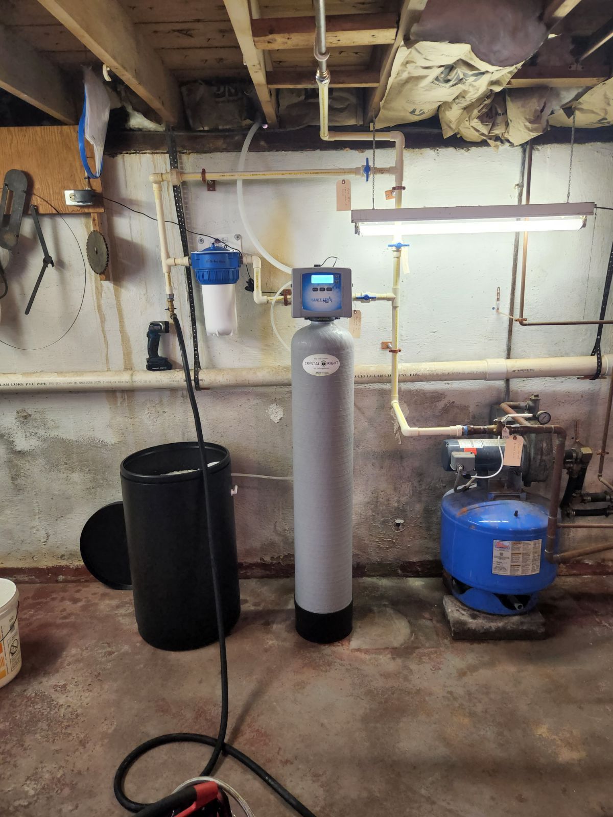 Water filtration system in a basement. Includes a tank, filter, pressure tank, and pipes against the wall.