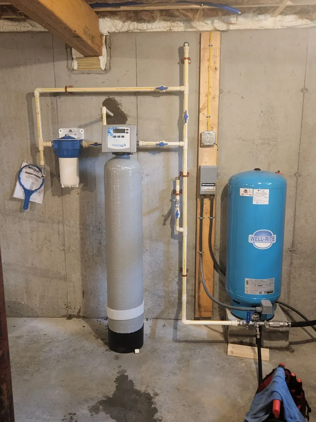 Water filtration system in a basement. Includes a blue tank, tall silver tank, and white filter, connected by beige pipes.