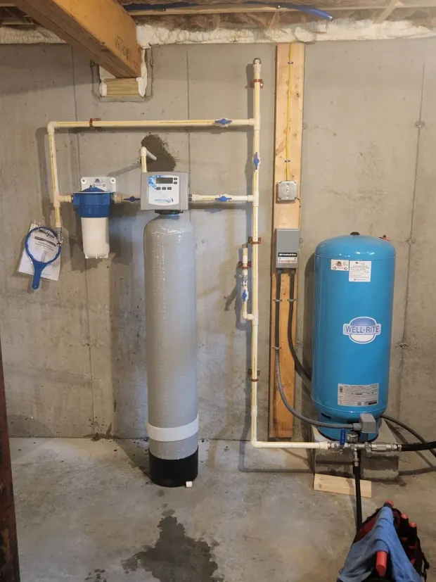 Water filtration system in a basement.  Includes a blue tank, tall silver tank, and white filter, connected by beige pipes.