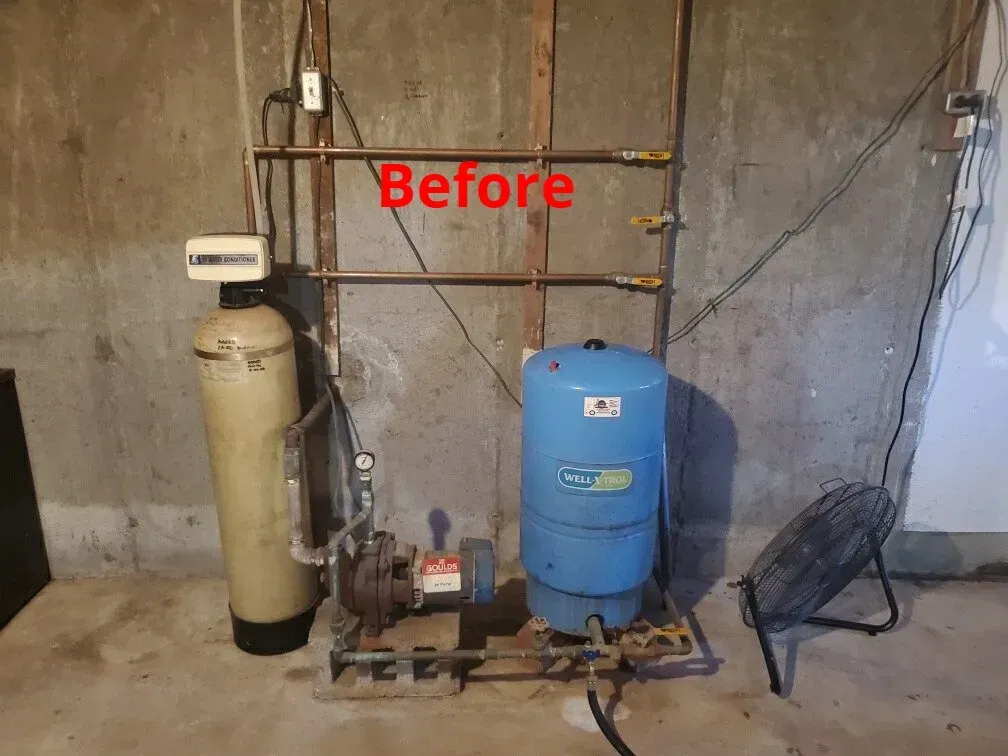 Water filtration and pressure tank setup against a concrete wall. Before image with pipes and a fan.