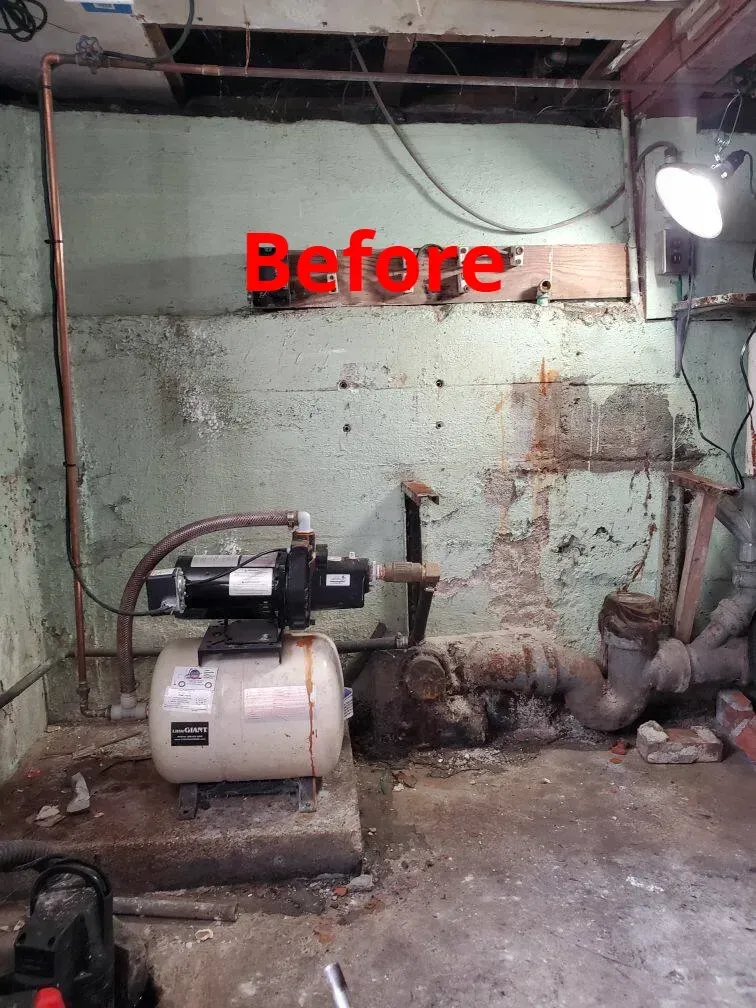 Basement water pump setup before renovation, with copper pipes, a white tank, and stained walls.
