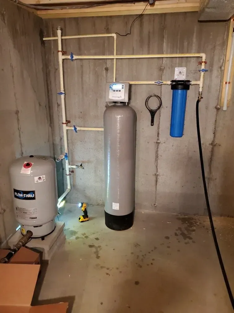 Plumbing in a basement: water softener, filter, pressure tank, and pipes on a concrete wall.