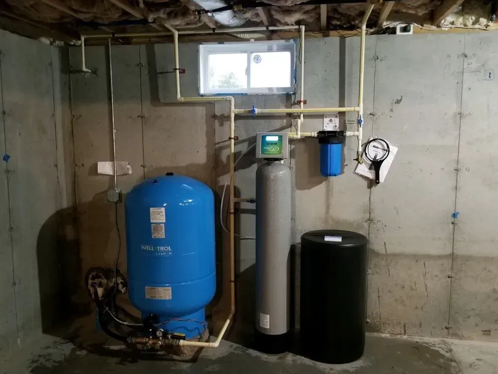 Water treatment system with blue pressure tank, filter, and black tank, installed against a concrete wall.