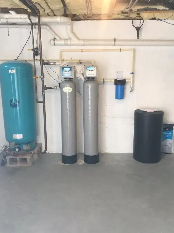 Water filtration system in a basement: blue tank, two gray tanks, blue filter, black tank, pipes against white wall.