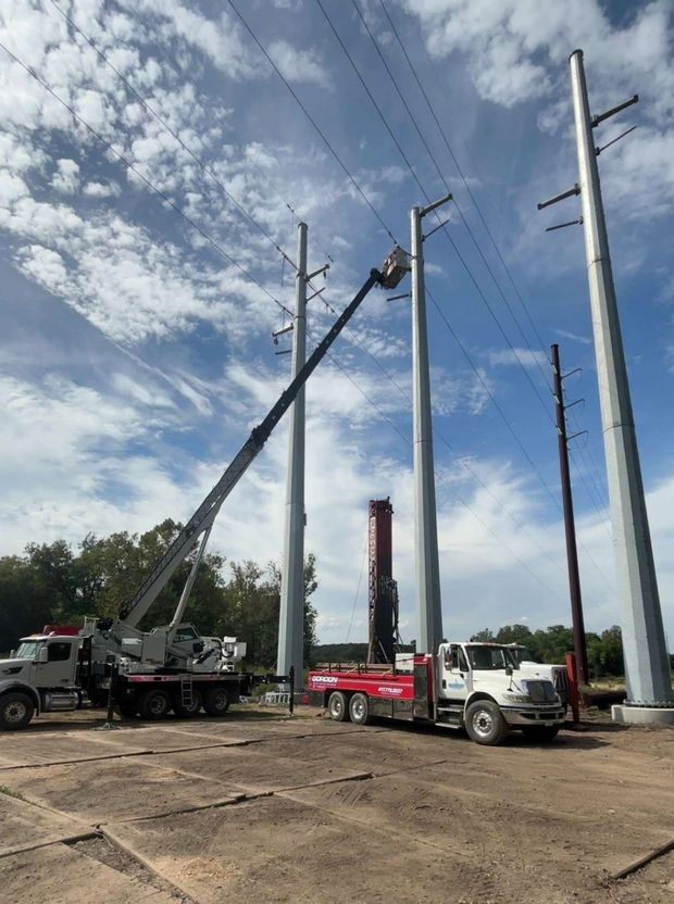 Construction trucks lifting utility poles in a dirt lot under a partly cloudy sky