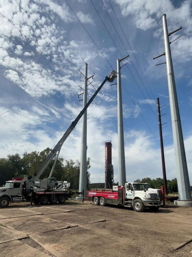 Construction trucks lifting utility poles in a dirt lot under a partly cloudy sky