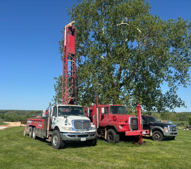 Three service trucks and a drilling rig parked on grass beside a tree under a clear blue sky.