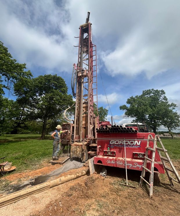 Drilling rig at a muddy worksite beside a red machine and trees under a cloudy sky