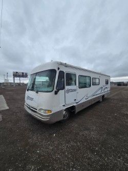 A white and tan motorhome parked on a gravel lot under a cloudy sky.
