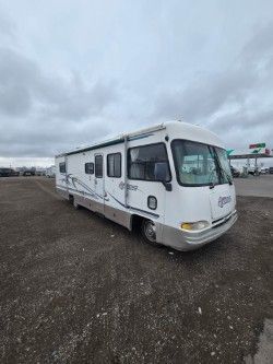 A white and tan Class A motorhome parked on a gravel lot under a cloudy sky.