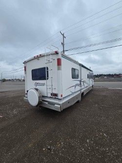 A white recreational vehicle parked on a gravel lot under a cloudy sky, featuring a rear ladder and spare tire cover.