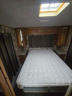 A queen bed with a white mattress inside a wood-paneled camper with a ceiling skylight and storage cabinets.