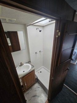 A view into a small RV bathroom featuring a white sink vanity, a shower stall with a skylight, and a wood-paneled door.
