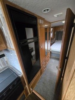 Interior view of an RV corridor showing a refrigerator, wood cabinetry, carpeted floor, and a bedroom in the background.