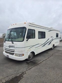 A white Class A motorhome with grey and black graphic accents parked on an asphalt lot under a cloudy sky.