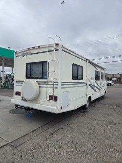 A white recreational vehicle parked at a gas station, seen from the rear angle, with a mounted ladder and spare tire.