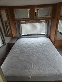 A queen-sized bed with a white quilted mattress pad, centered beneath wooden cabinets and a window with pleated blinds.