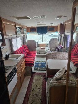 Interior of an RV with wood cabinetry, a red patterned sofa, a small dining table, and a view through the front window.