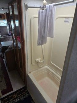 An RV bathroom featuring a white shower stall with a curtain rod and a hanging white towel, adjacent to a wooden cabinet.