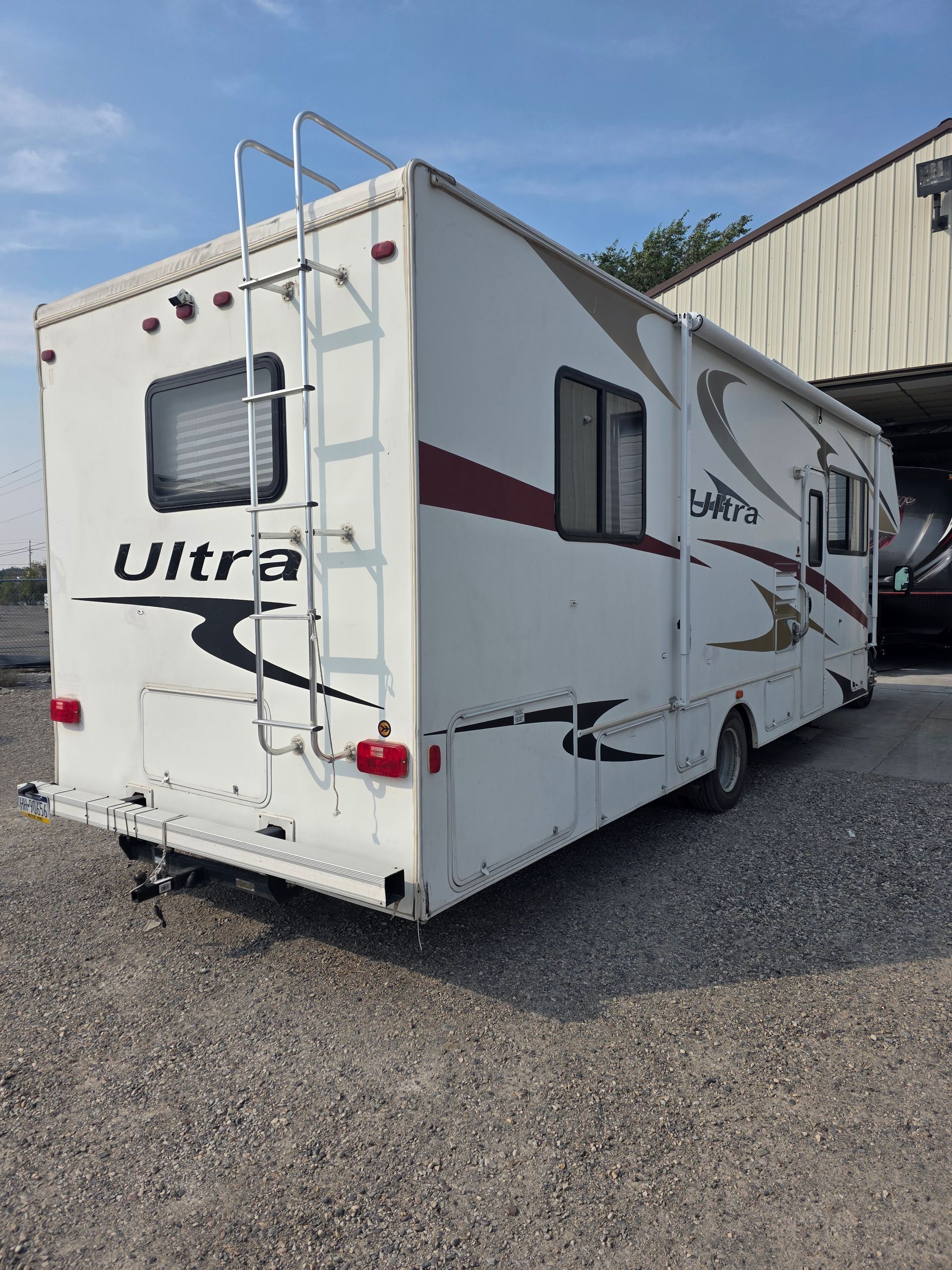White RV parked on gravel, with ladder on the back.