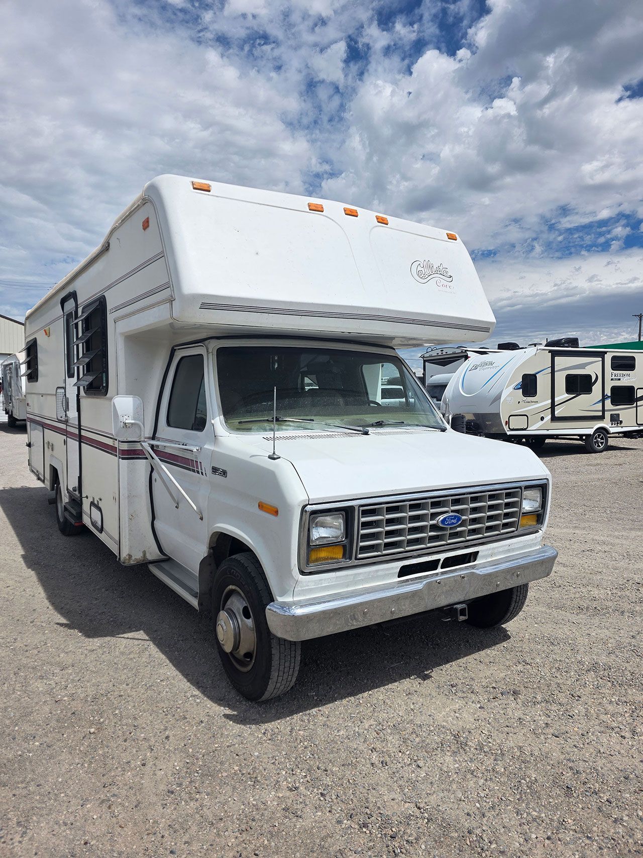 White RV parked on gravel lot. Overhead storage, front cab, and side windows visible. Blue sky background.