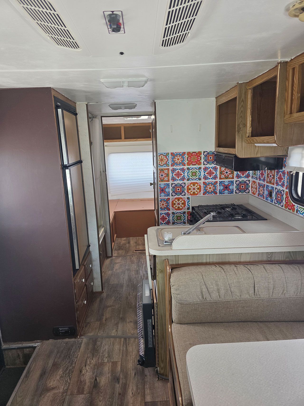 Interior of a camper, showing kitchen area with stove, fridge, and seating. Wooden cabinets and colorful tiled backsplash are visible.