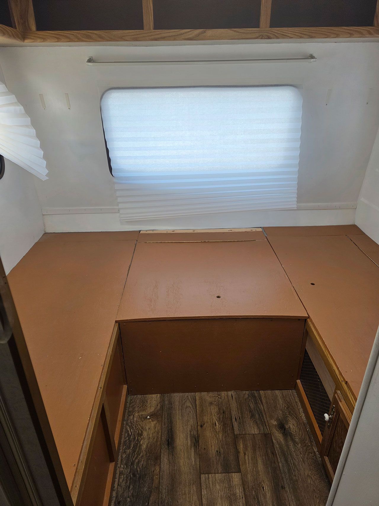 Interior of a small trailer with a bed platform and window. Brown platform and cabinets with a wood-look floor.