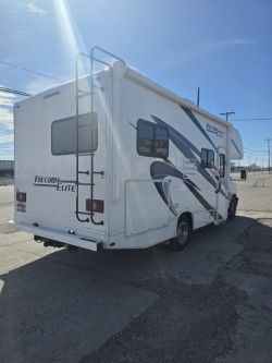 A white Class C motorhome with blue and gray side graphics, viewed from the rear passenger side in a sunny parking lot.