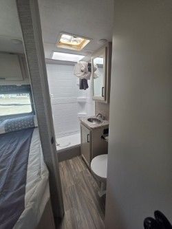 A view from a doorway into a small RV bathroom with a shower, a sink vanity, a toilet, and light wood-look flooring.