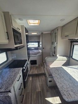 Interior view of a camper with a kitchen, dining area, and a bed in the background, featuring light wood-toned cabinetry.