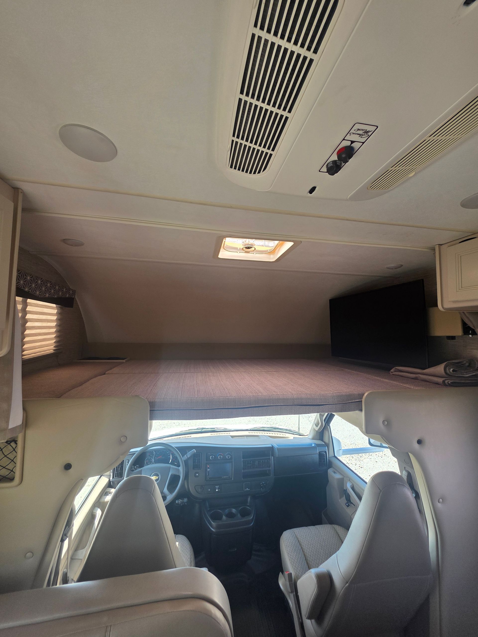 Interior view of an RV cabin, showing two front seats, a dashboard, and a raised bunk area with a television above.