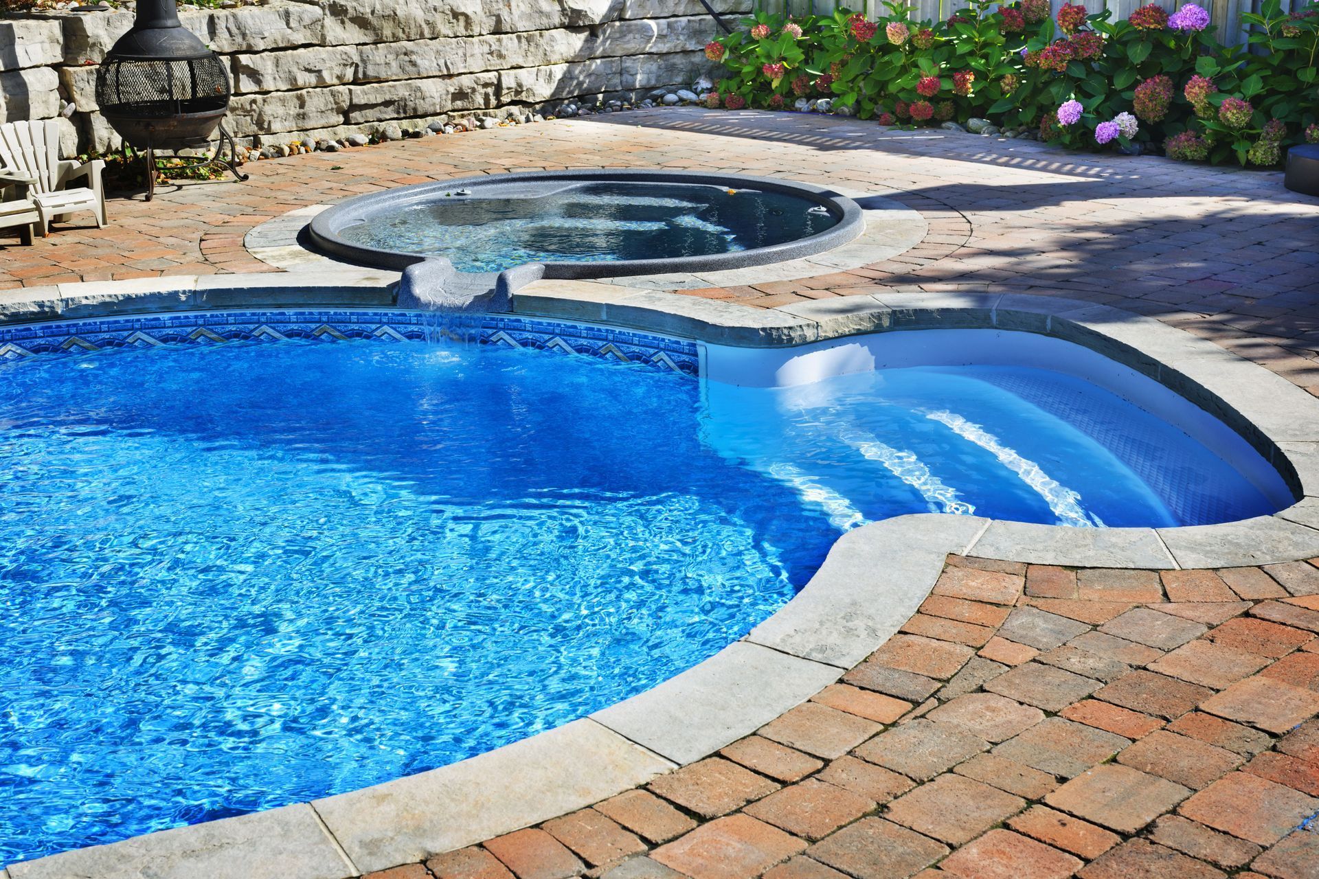 Blue pool with brickwork surround, hot tub, and stone wall backdrop.