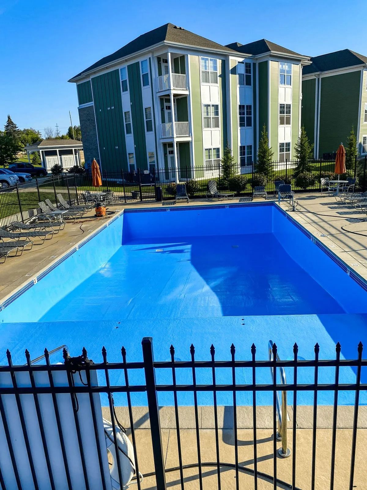 Empty blue swimming pool behind a black fence, with green apartment buildings in the background.