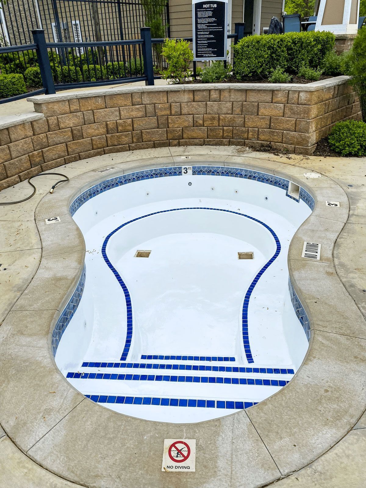 Empty, kidney-shaped hot tub with blue tile trim, surrounded by concrete. Brick wall and greenery in the background.