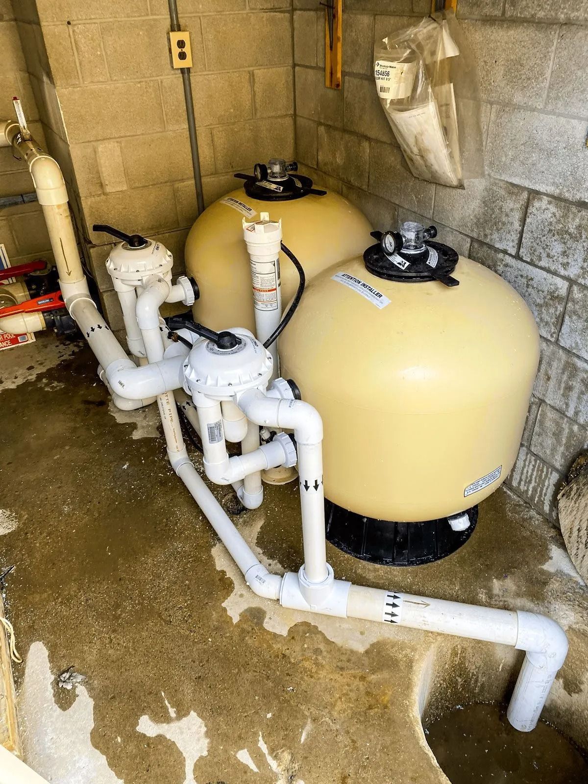 Pool filter system with sand filters and PVC pipes in a concrete enclosure.