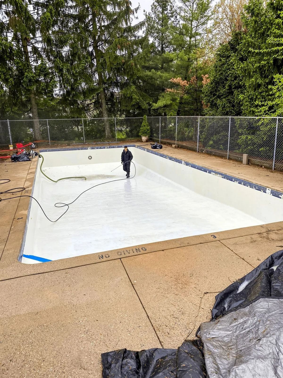Person painting the interior of an empty rectangular swimming pool white. Trees and a fence in the background.