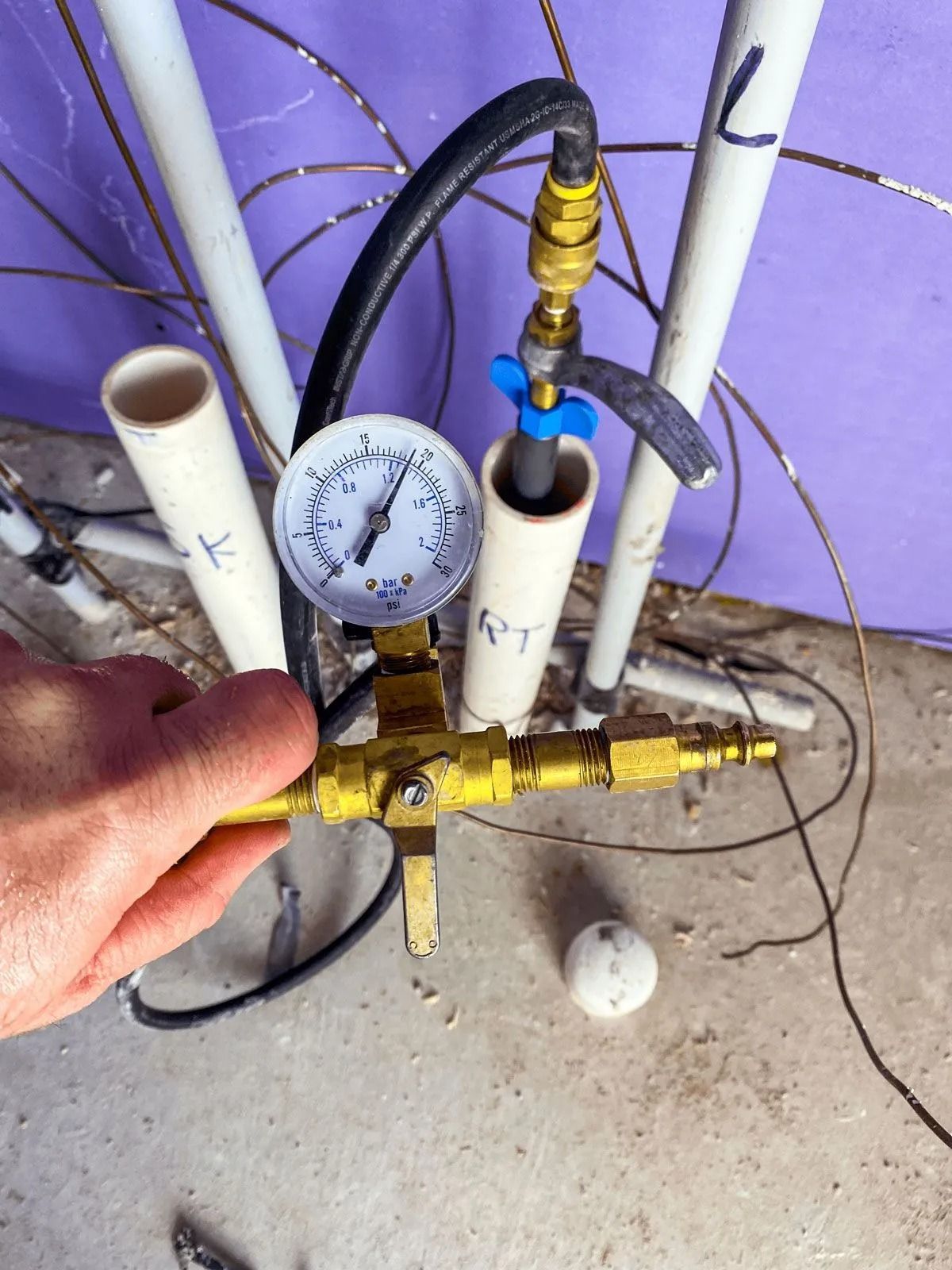 A hand holds a brass tool connected to a pressure gauge. White pipes and wires are in the background, likely a construction site.