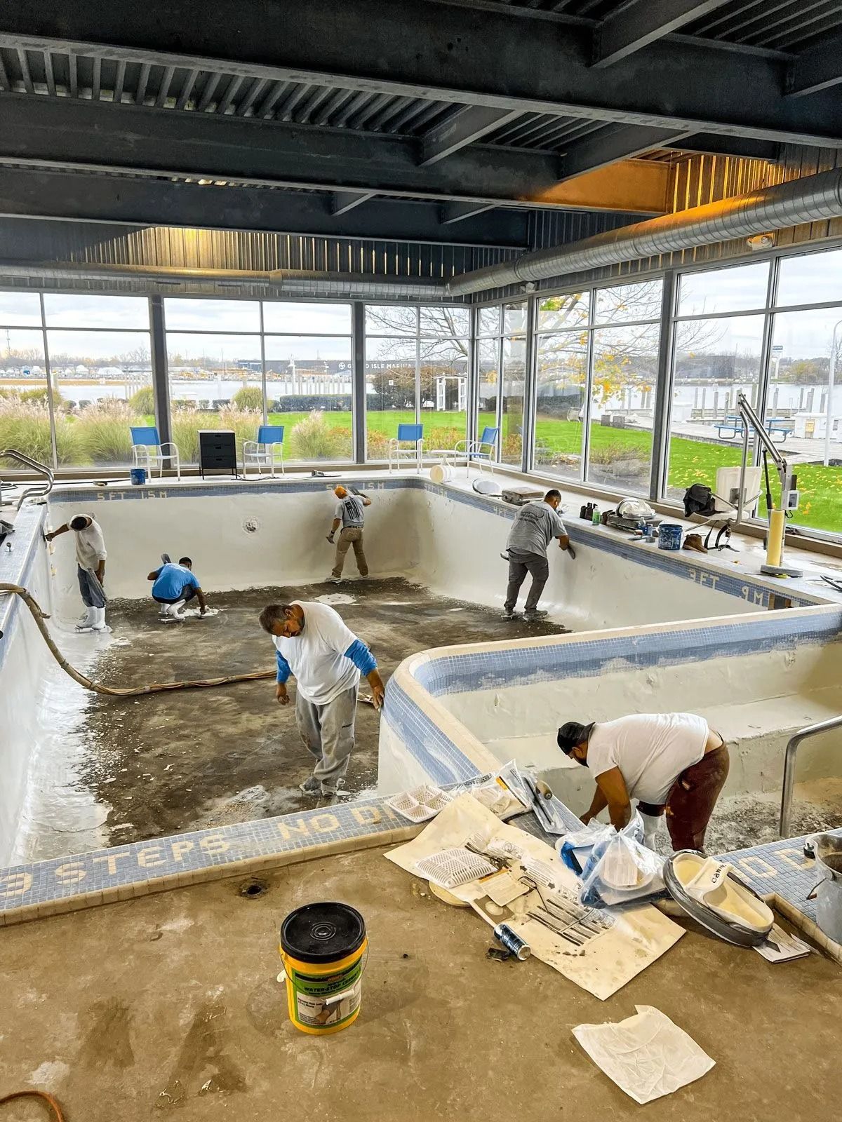 Workers renovating an indoor pool with exposed beams and large windows overlooking a body of water.