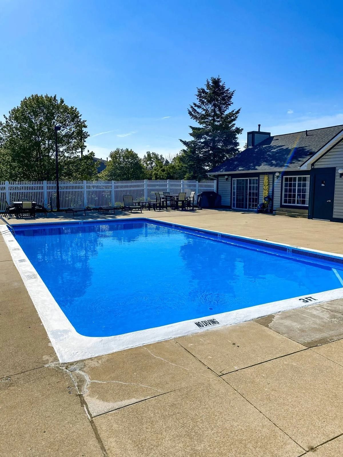 Rectangular swimming pool filled with bright blue water on a sunny day. White fence and building visible.