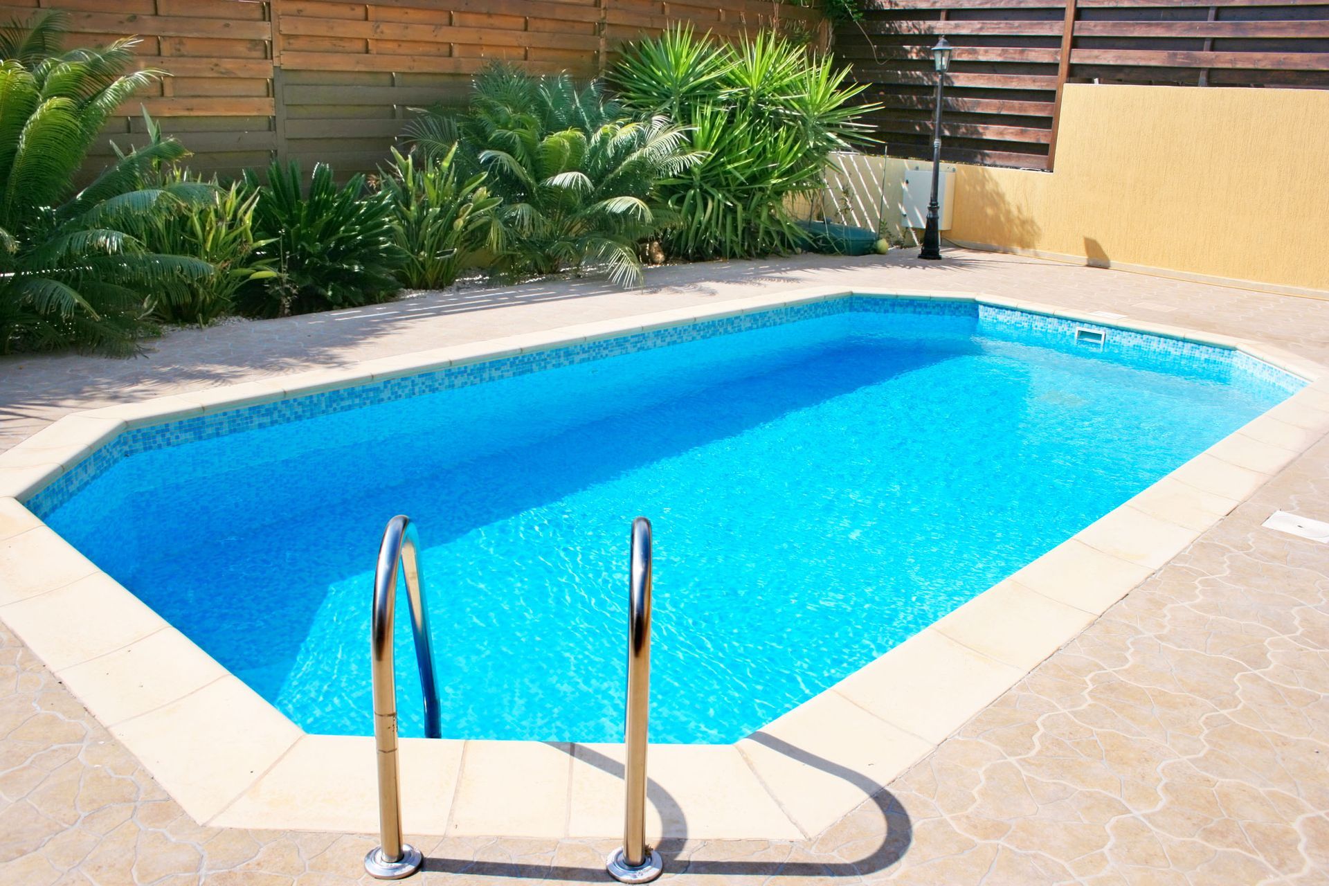 Swimming pool with blue water and chrome ladder surrounded by beige paving stones and greenery.