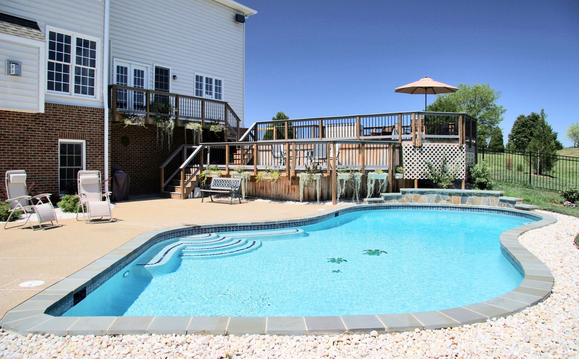 Swimming pool next to a two-story house with a wooden deck. The sky is blue and there are two lounge chairs visible.