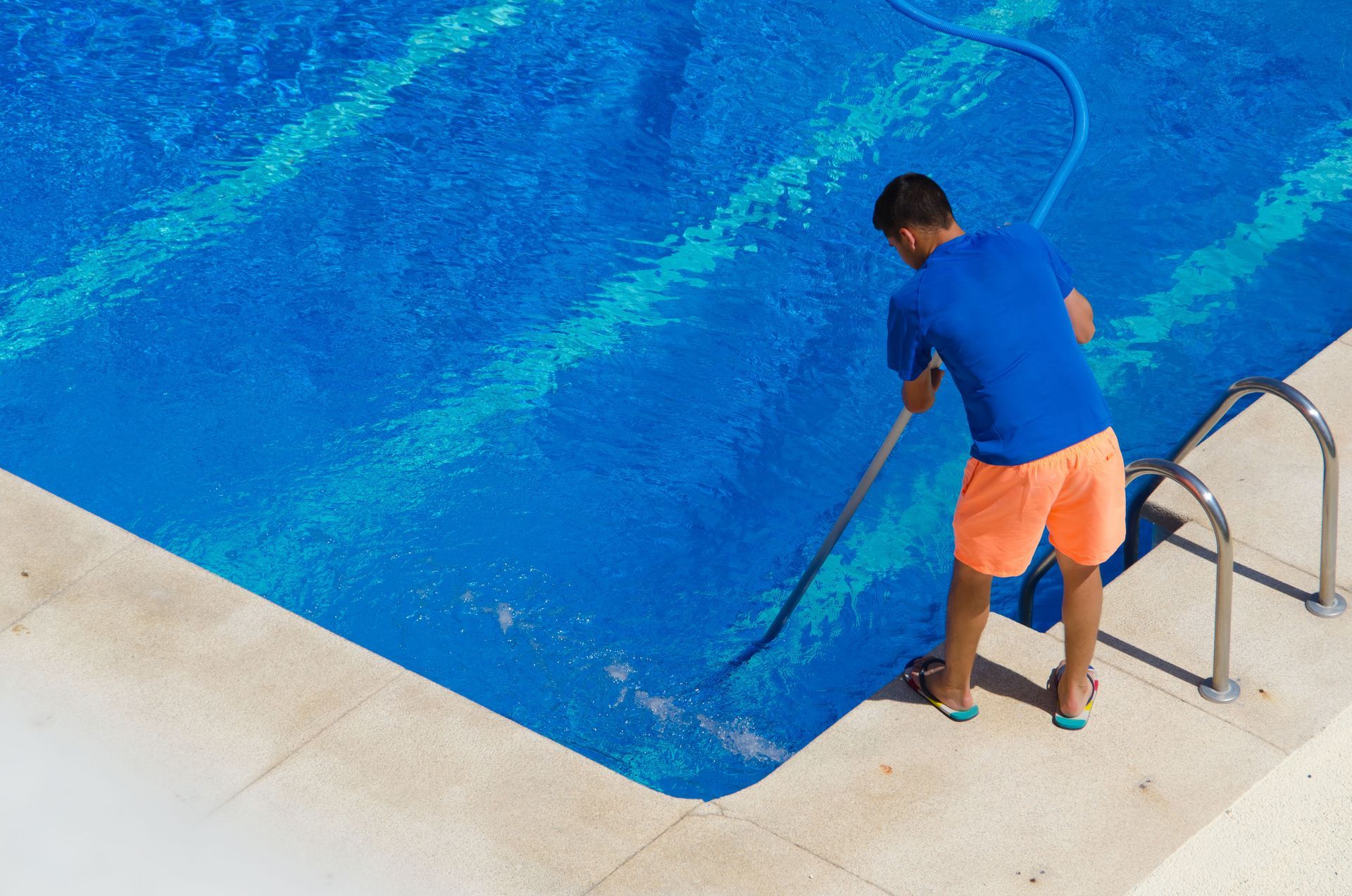 Person in blue shirt and orange shorts cleaning a bright blue pool with a long-handled vacuum.