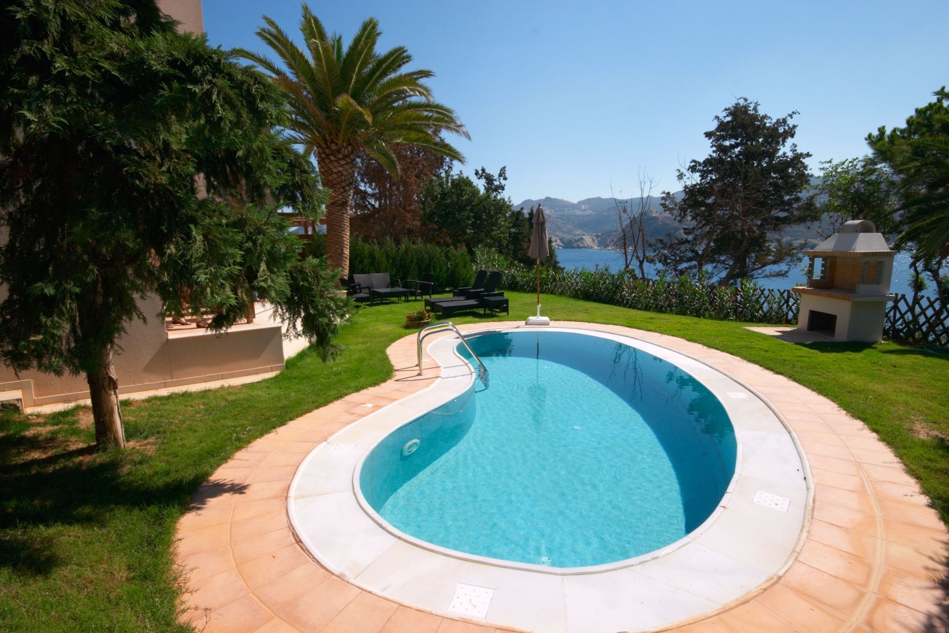Pool with blue water surrounded by lawn, trees, and overlooking a bay. Sunny day.