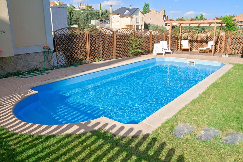 Blue rectangular swimming pool in a backyard surrounded by grass, a fence, and lounge chairs.