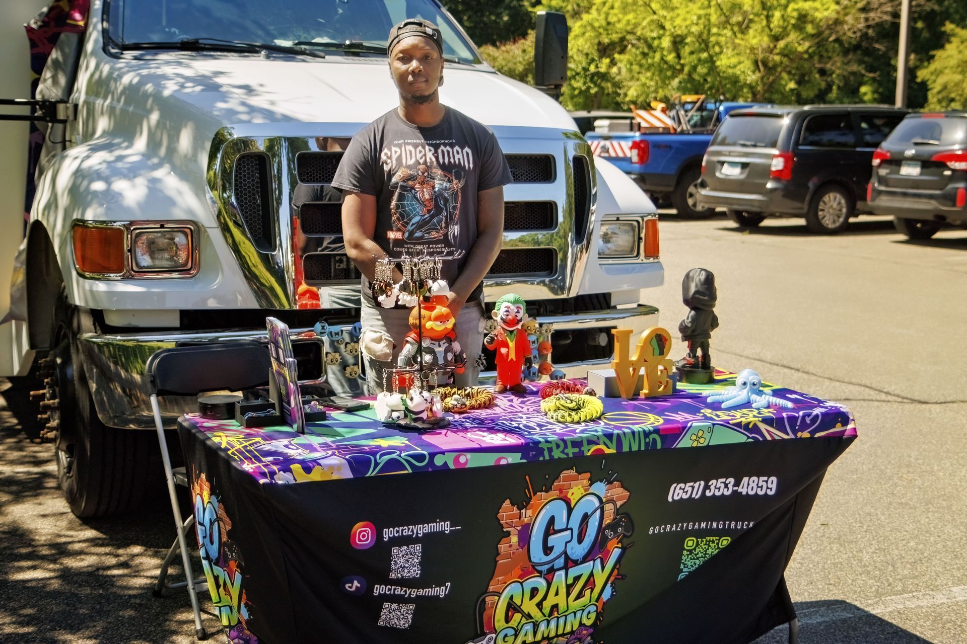 Man standing behind a colorful vendor table in front of a white pickup truck outdoors